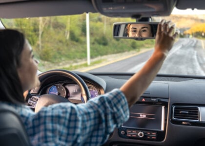 Attractive young woman driving a car, view from inside. A woman driver touches the mirror in the car. The concept of a long road, traveling by car.