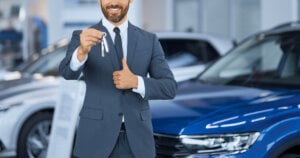 Close up of bearded man in suit showing keys from new car on camera while standing at auto center. Happy male owner of new transport showing thumb up. Expensive purchase of businessman.