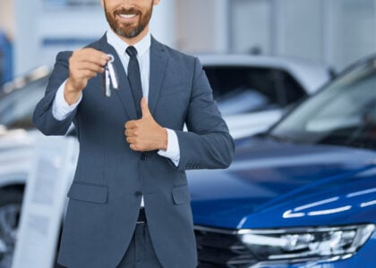 Close up of bearded man in suit showing keys from new car on camera while standing at auto center. Happy male owner of new transport showing thumb up. Expensive purchase of businessman.