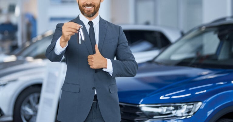 Close up of bearded man in suit showing keys from new car on camera while standing at auto center. Happy male owner of new transport showing thumb up. Expensive purchase of businessman.
