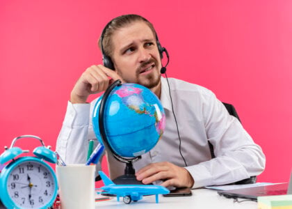 handsome businessman in white shirt with headphones touching globe looking aside confused sitting at the table in offise over pink background