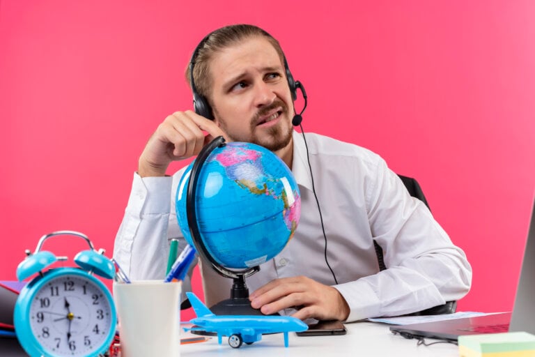 handsome businessman in white shirt with headphones touching globe looking aside confused sitting at the table in offise over pink background